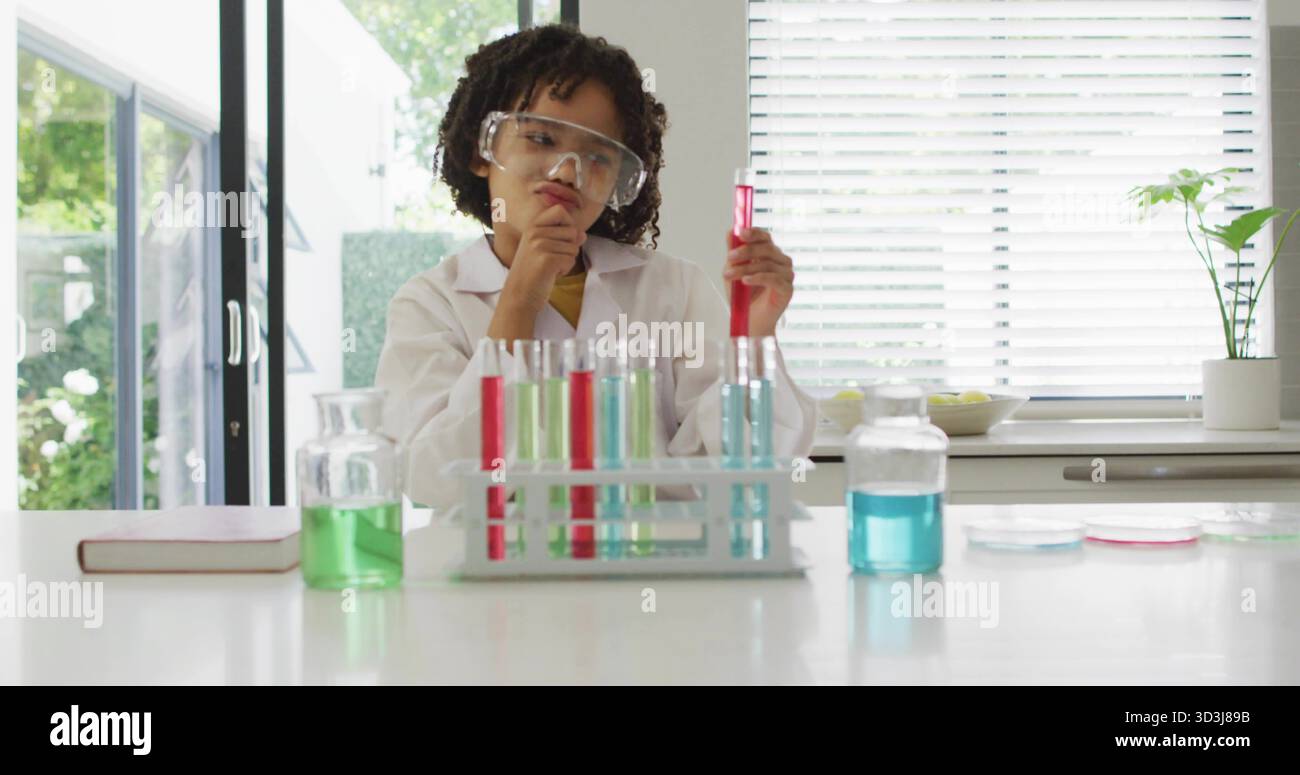 Enfant en blouse de laboratoire examinant un tube à essai rouge sur le comptoir de cuisine, avec un portoir pour tubes à essai Banque D'Images