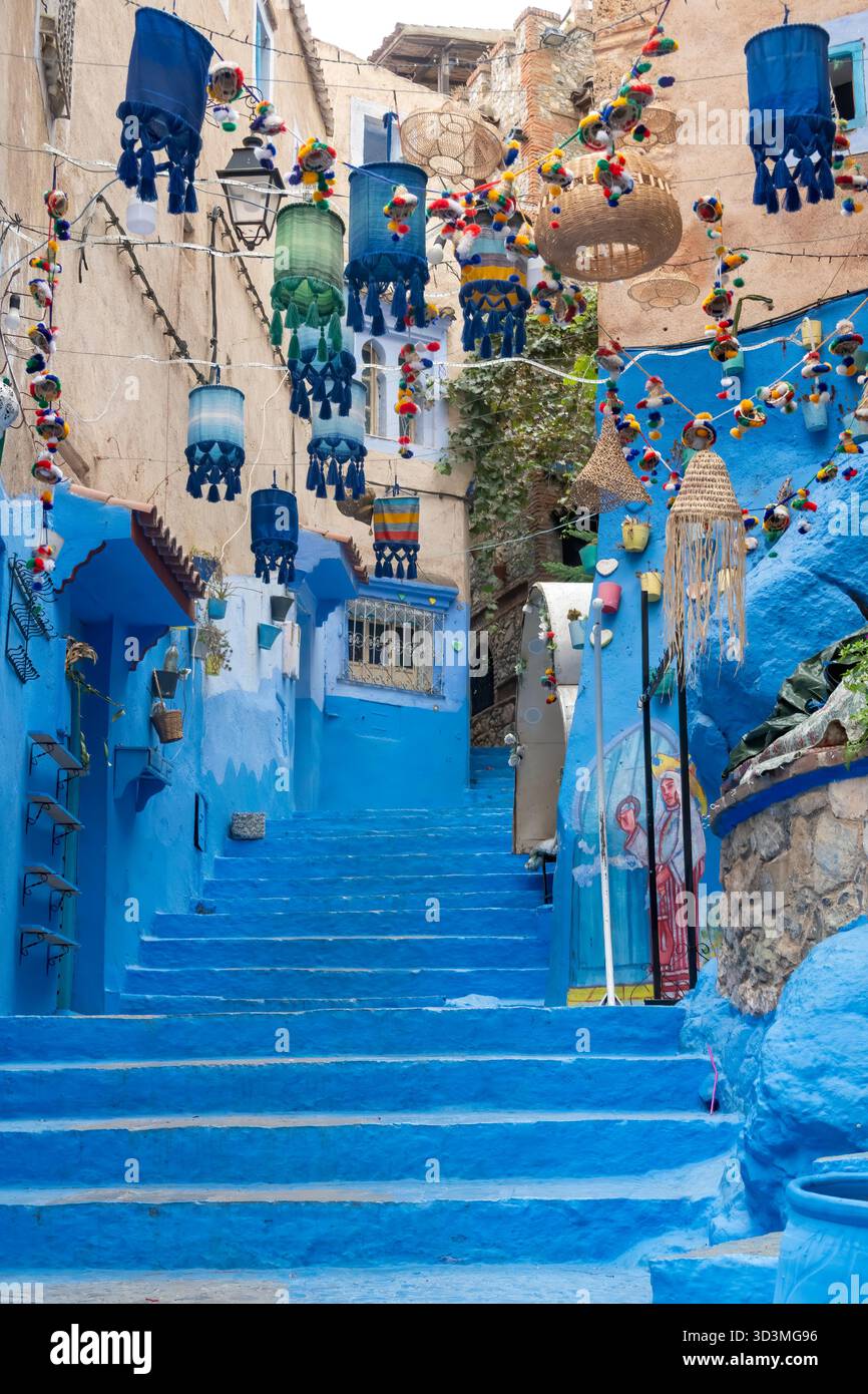 Une ruelle escarpée, bleue et blanche à Chefchaouen, au Maroc, est fortement décorée. De nombreuses lanternes marocaines colorées pendent au-dessus de la tête, créant un et unique Banque D'Images