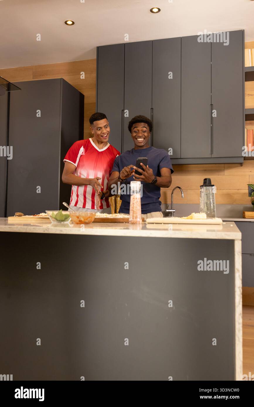 Rire divers amis masculins vérifiant smartphone dans la cuisine à la maison, avec planche à découper et bols Banque D'Images