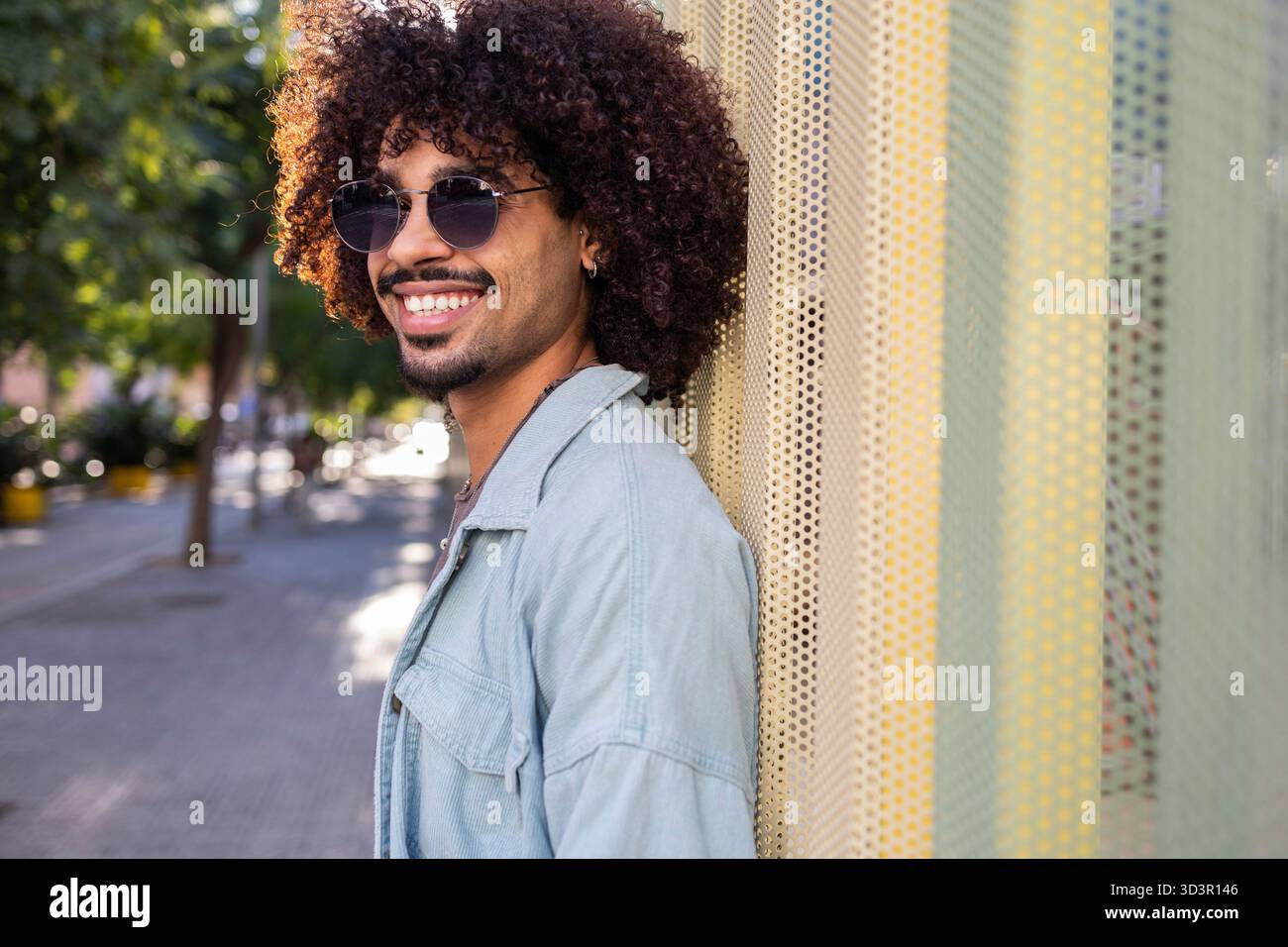 Un homme élégant avec des cheveux bouclés et des lunettes de soleil profite d'une journée ensoleillée dans la ville. Il s'appuie contre un mur texturé, exsudant confiance et cha urbain moderne Banque D'Images