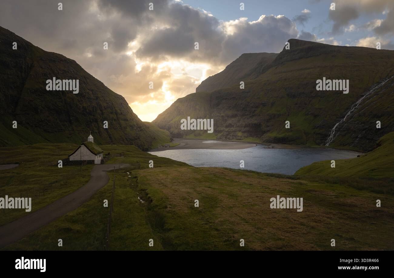 Vue imprenable sur une vallée sereine dans les îles Féroé, avec une église pittoresque et une rivière tranquille entourée de collines verdoyantes, sous un Banque D'Images