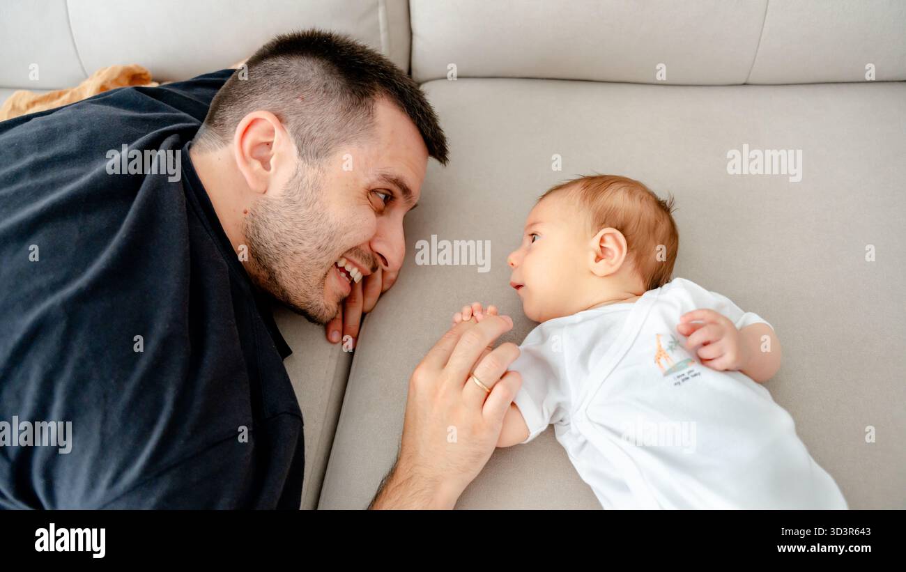 Un père allongé à côté de son nouveau-né sur un canapé confortable, partageant un moment tendre. Le bébé regarde curieusement le père, symbolisant la beauté du nouveau pare Banque D'Images