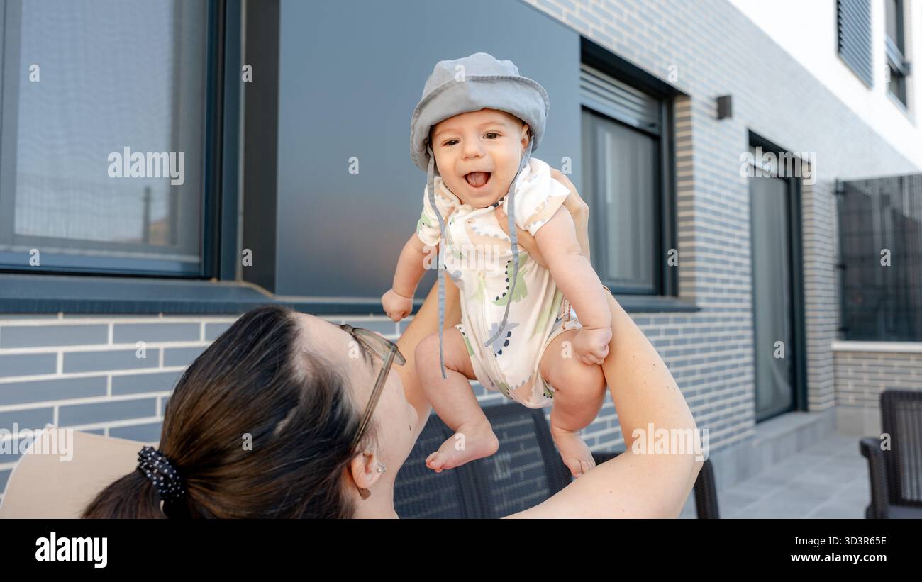 Une mère joyeuse soulève son bébé heureux dans l'air à l'extérieur d'un bâtiment moderne, mettant en valeur un moment de lien familial et de développement de la petite enfance Banque D'Images