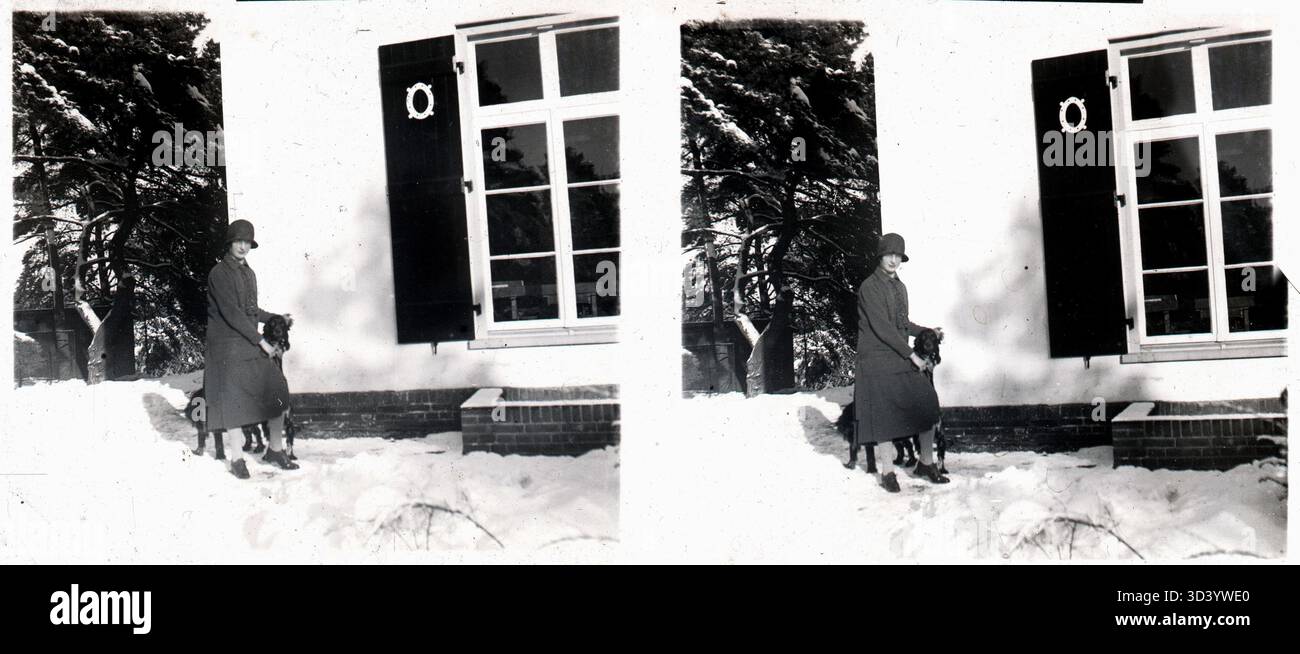 Une femme portant un chapeau et un long manteau d'hiver se tient avec un chien dans la neige près d'une maison, vers 1900-1930. La photo fait partie d'une série de plaques de verre stéréoscopiques. Banque D'Images