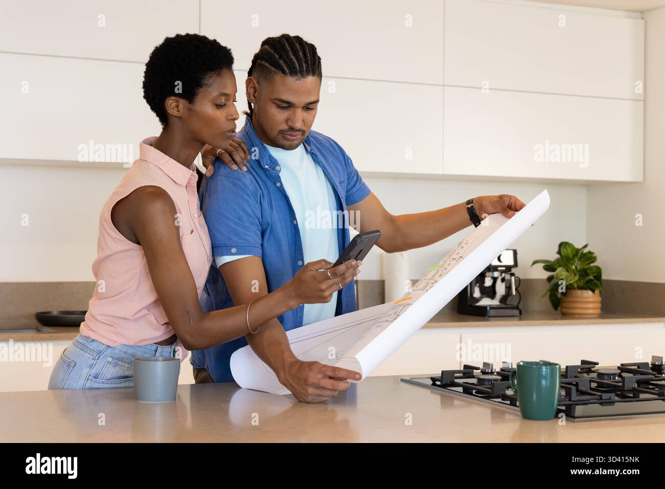 Examen de couple diversifié étudiant le plan de conception sur l'îlot de cuisine, avec smartphone et tasses Banque D'Images