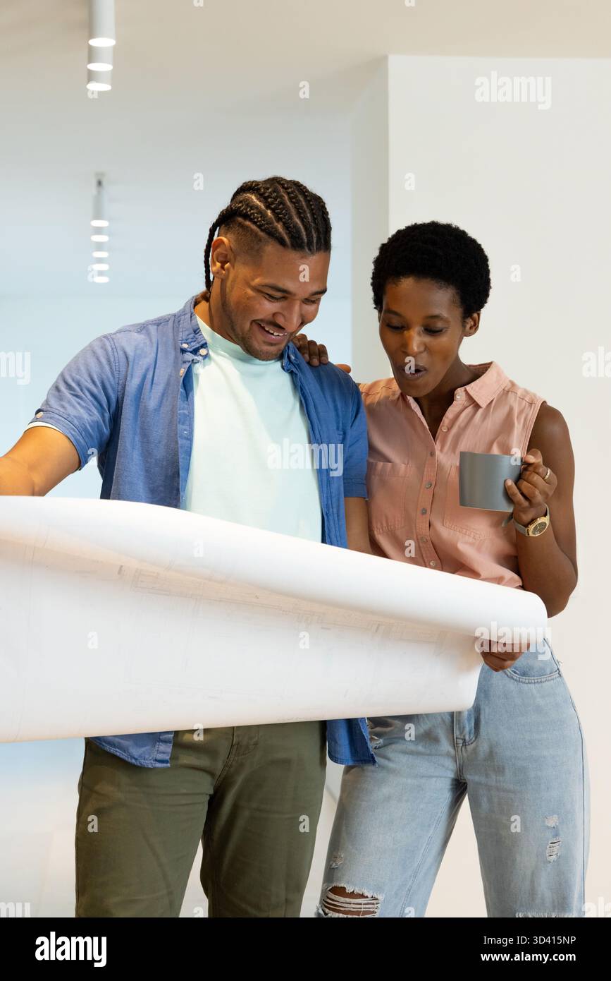 Couple diversifié déroulant le plan architectural dans un studio de design moderne, avec tasse à café Banque D'Images