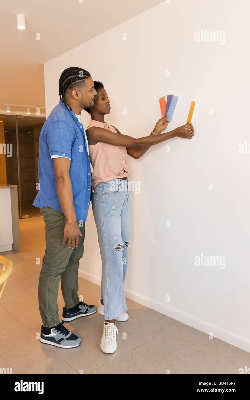 Comparer un couple diversifié tenant quatre échantillons de peinture contre le mur blanc dans la maison moderne, copier l'espace Banque D'Images