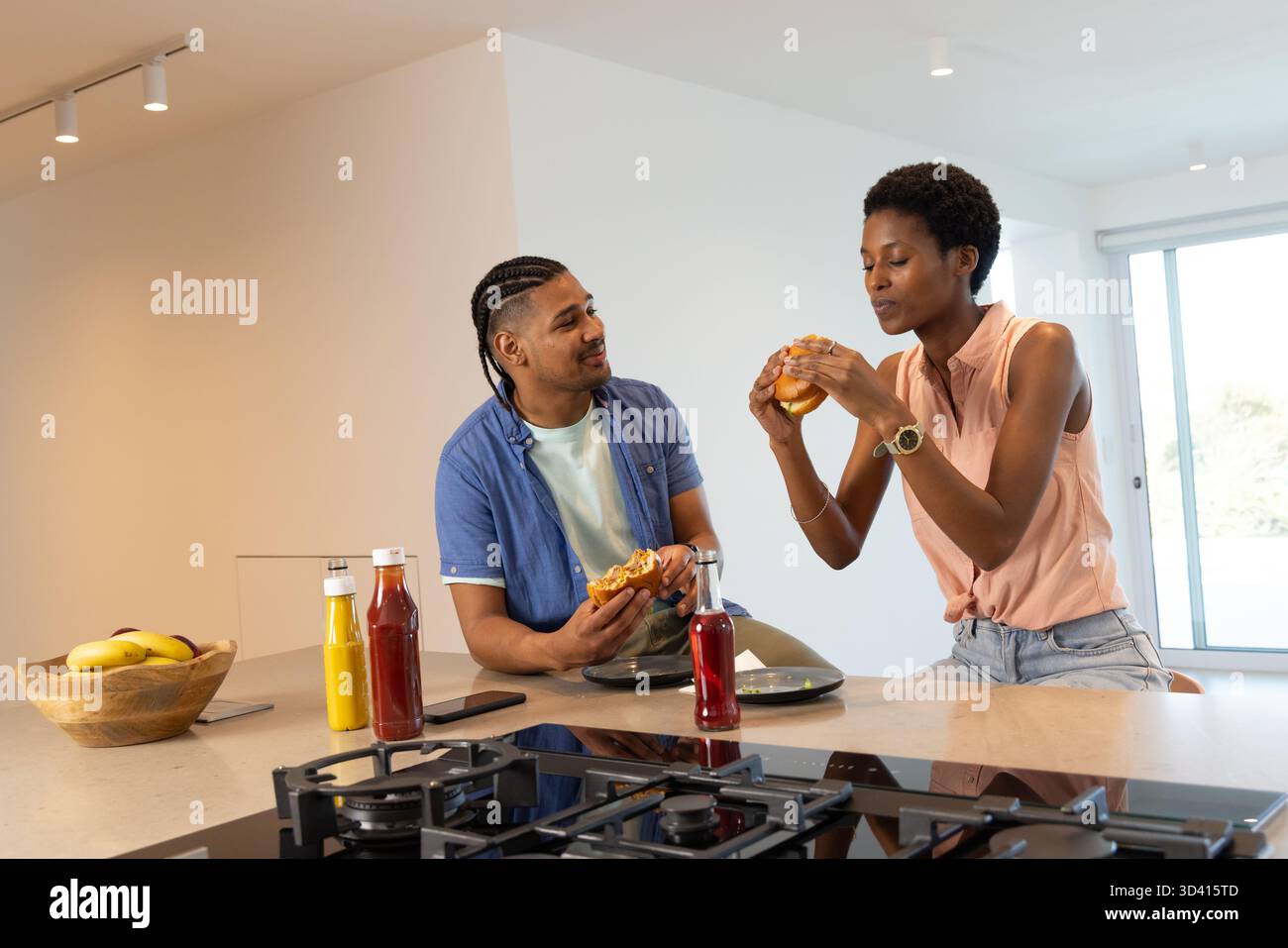 Couple diversifié mangeant des hamburgers à l'îlot de cuisine, avec bol de bananes et bouteilles de ketchup Banque D'Images