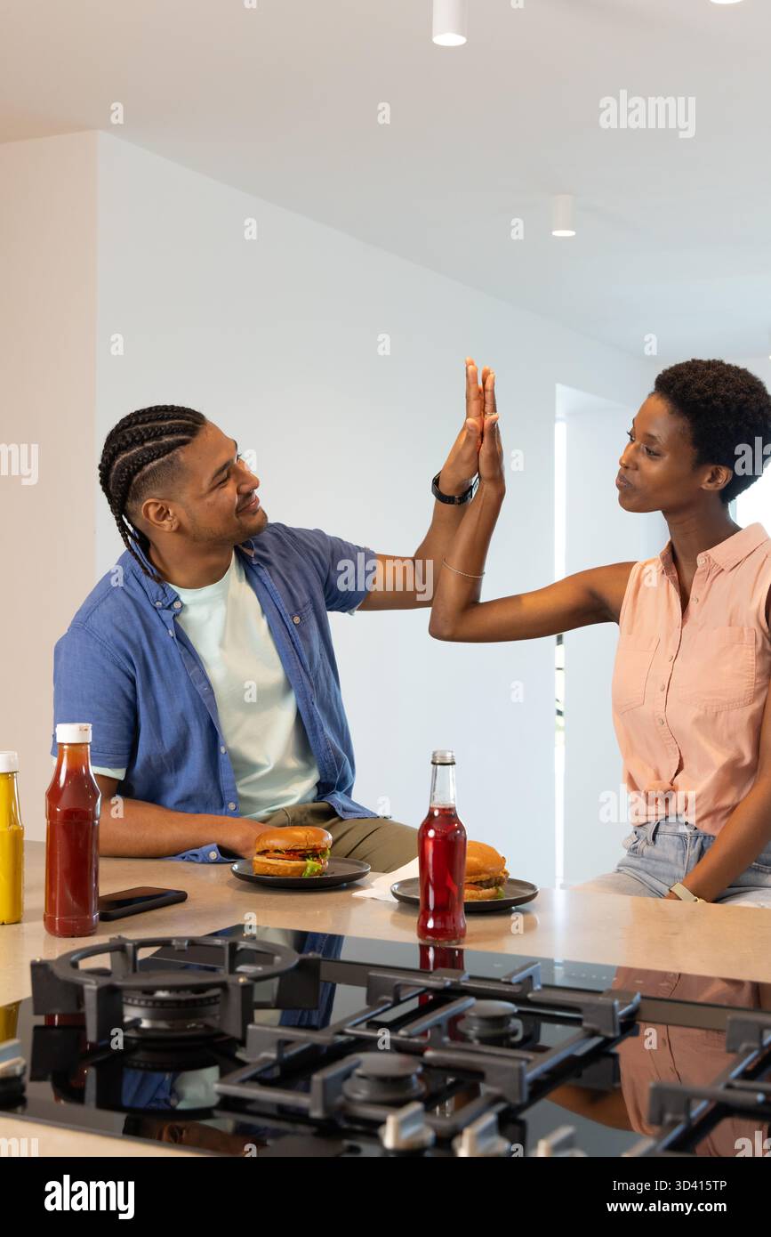 Des amis diversifiés mangeant des hamburgers sur l'îlot de cuisine moderne, avec smartphone et boissons rouges Banque D'Images