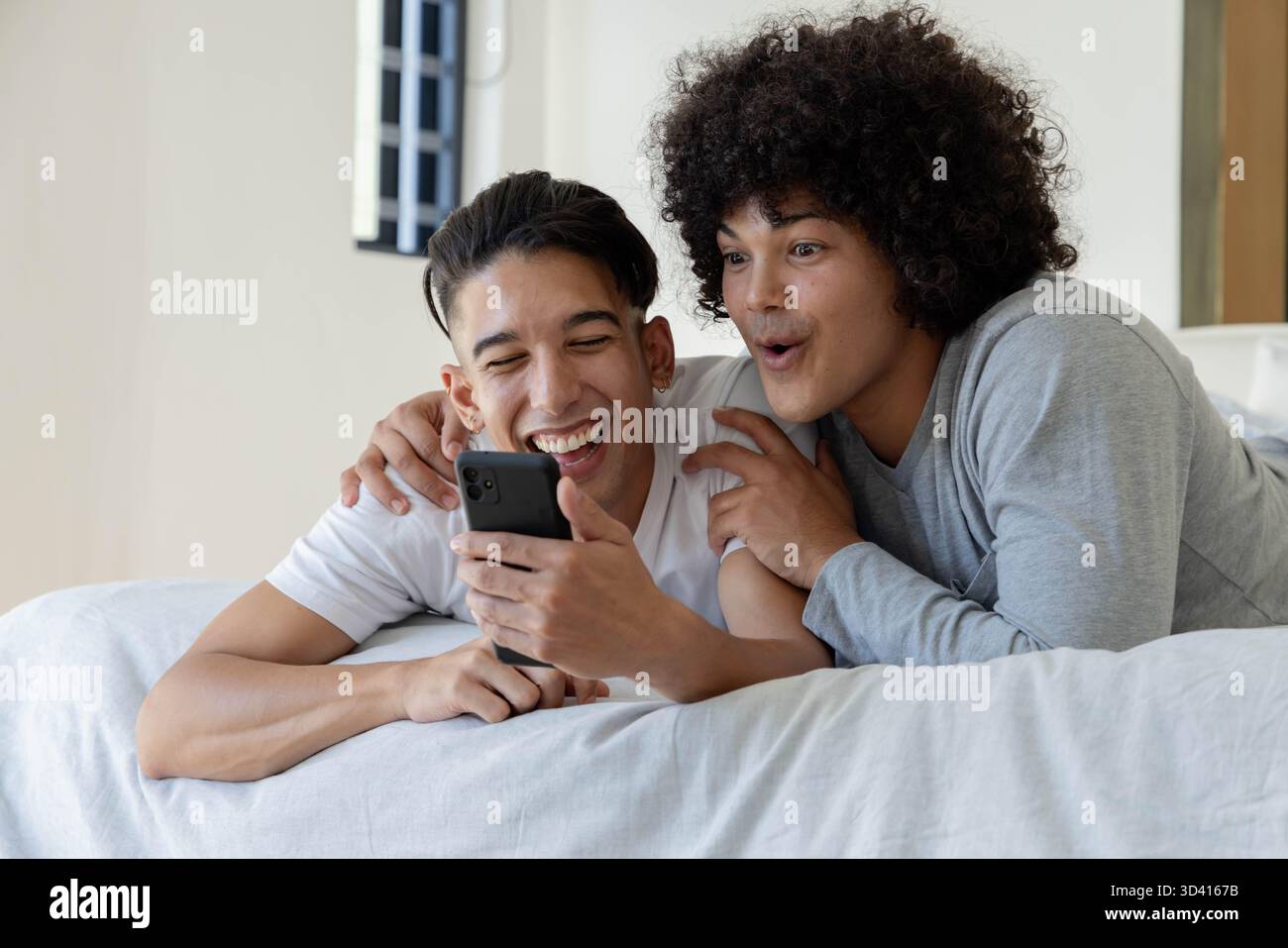 Rire divers amis masculins allongés sur le lit dans la chambre à coucher regardant smartphone, avec des draps blancs Banque D'Images