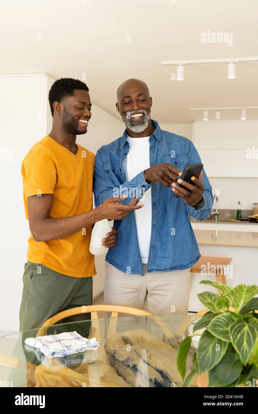 Sourire père afro-américain et fils nettoyant la cuisine ensemble, avec flacon pulvérisateur et smartphone Banque D'Images