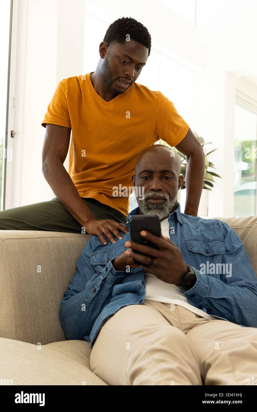 Père afro-américain et son fils à l'aide d'un smartphone sur un canapé beige dans le salon, avec plante, fenêtres Banque D'Images