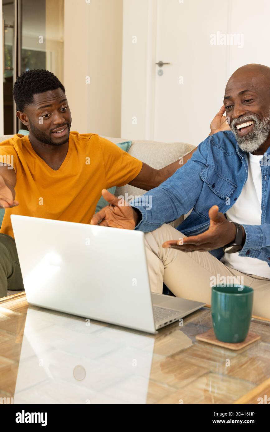 Rire père afro-américain et fils faisant des gestes vers l'ordinateur portable dans le salon, avec tasse verte Banque D'Images