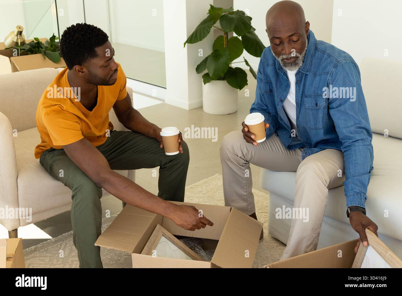 Déballage père afro-américain et fils ouvrant des boîtes dans le salon, avec tasses à café, cadres Banque D'Images