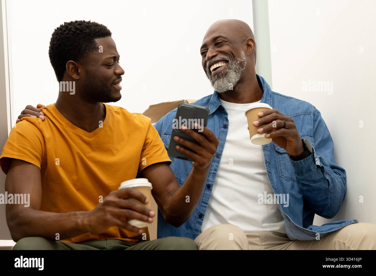 Assis père afro-américain et fils tenant des tasses à café sur le sol à la maison, avec smartphone Banque D'Images