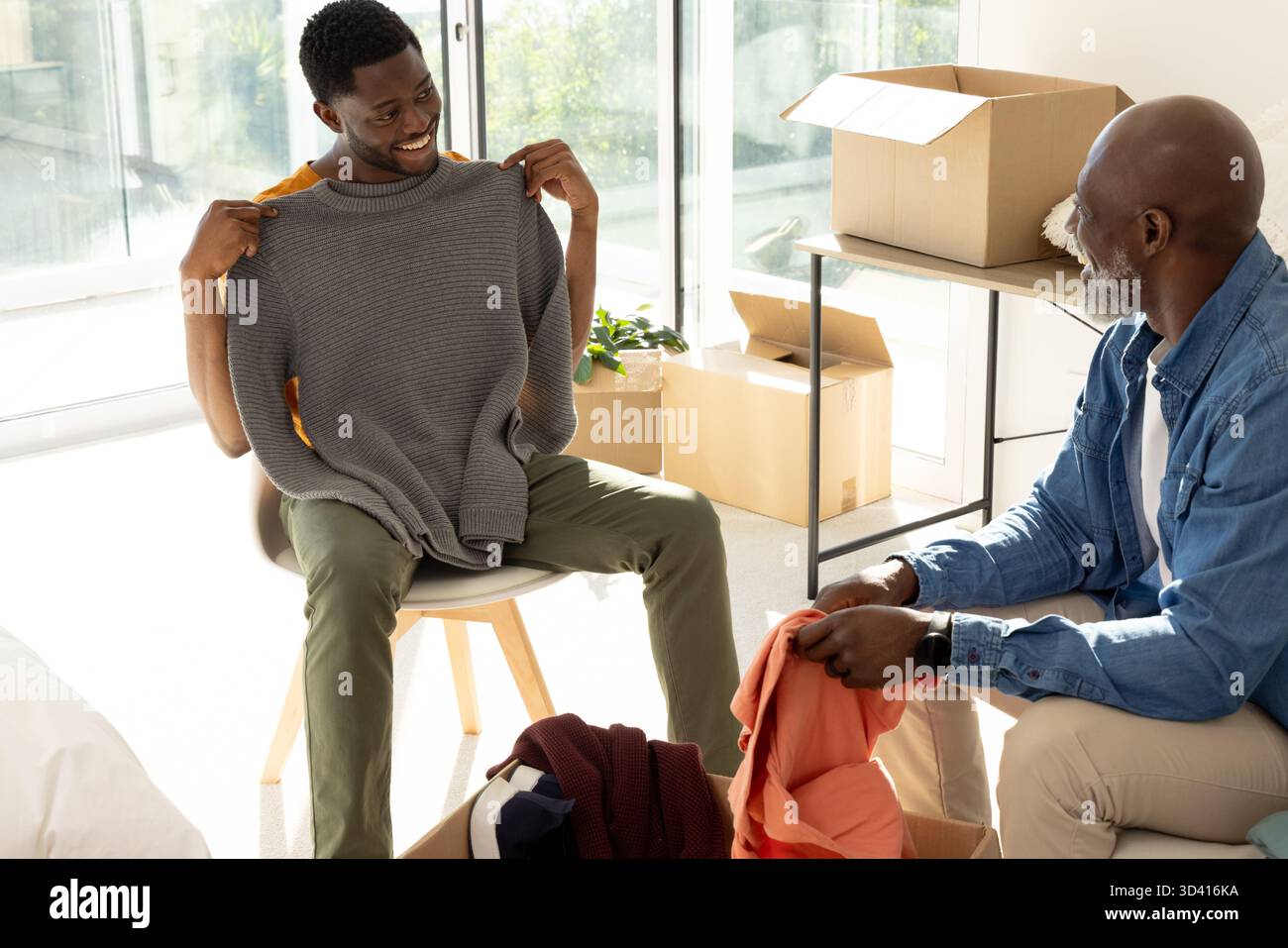 Examinant un père et un fils afro-américains triant des vêtements dans un salon ensoleillé, avec des boîtes mobiles Banque D'Images