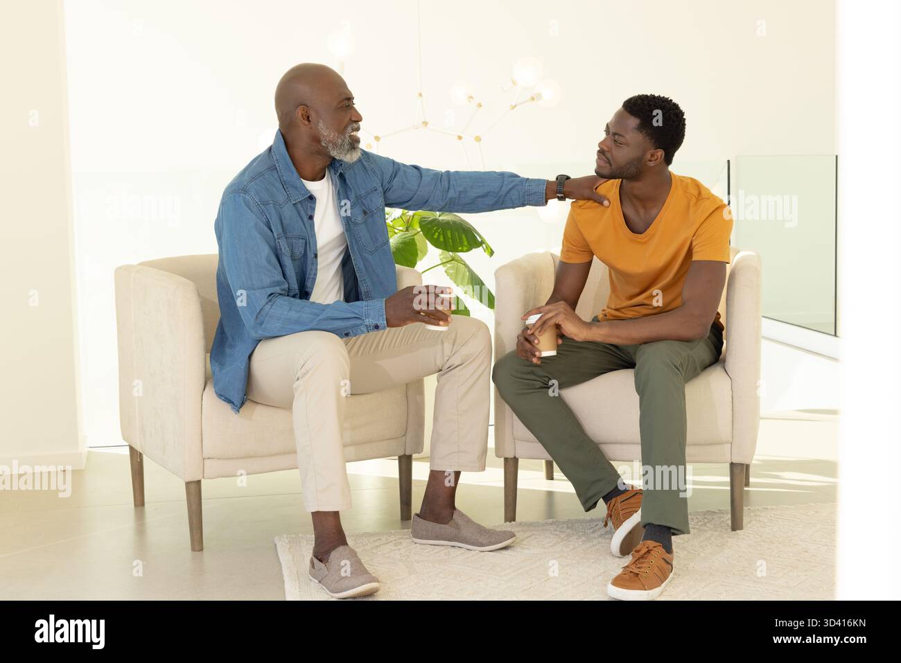 Bavardant père et fils afro-américains assis sur des chaises beiges dans le salon, tenant des tasses blanches Banque D'Images