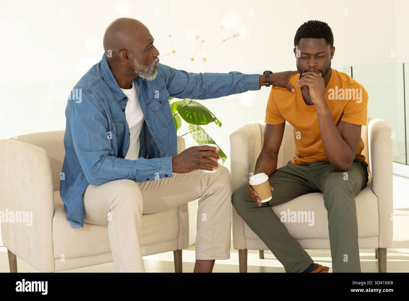 Soutien père afro-américain et fils assis dans des fauteuils dans le salon, avec tasses à café Banque D'Images
