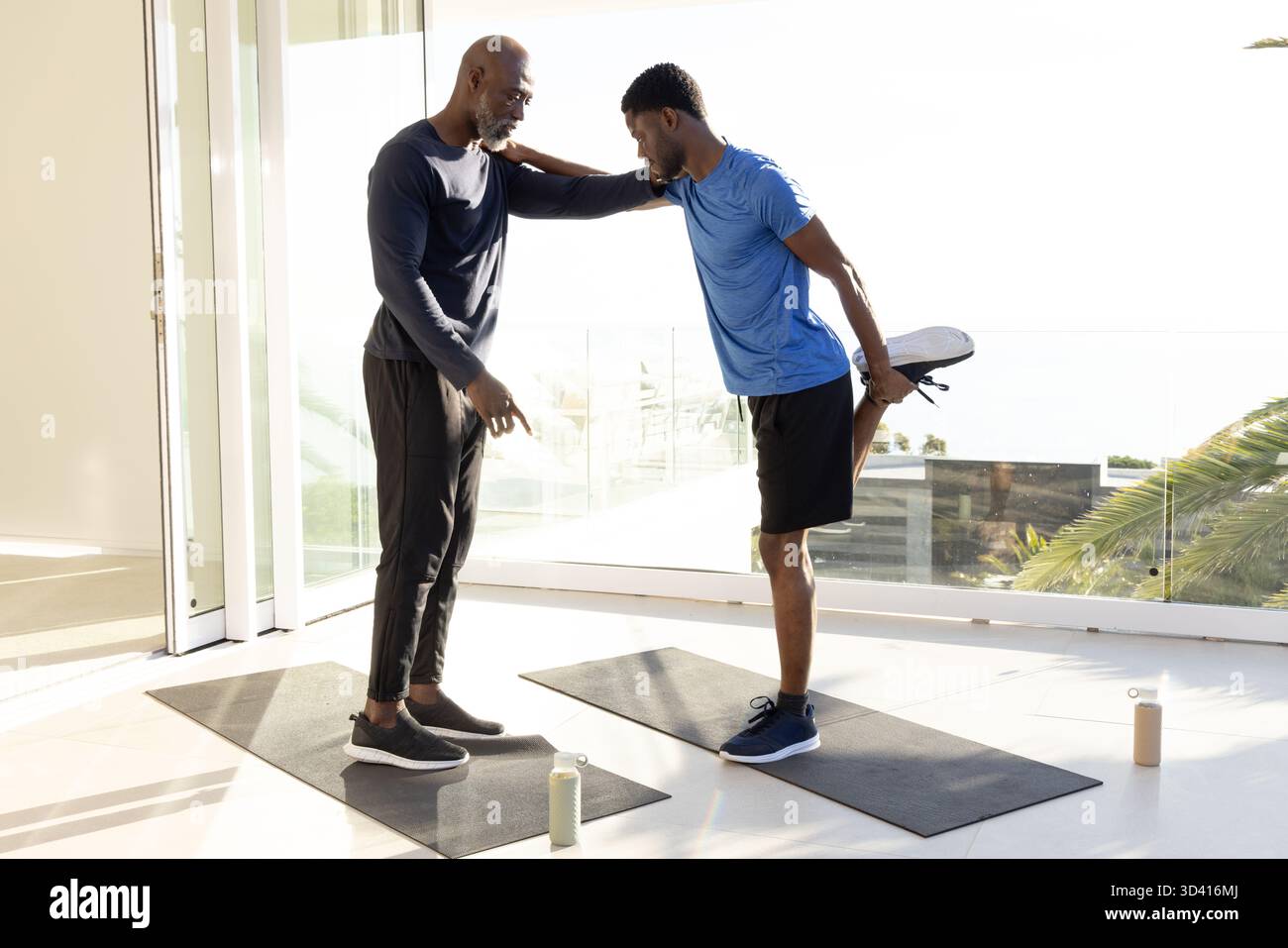 Entraîneur afro-américain et client faisant étirer les jambes sur des tapis noirs dans le gymnase, avec des bouteilles d'eau Banque D'Images
