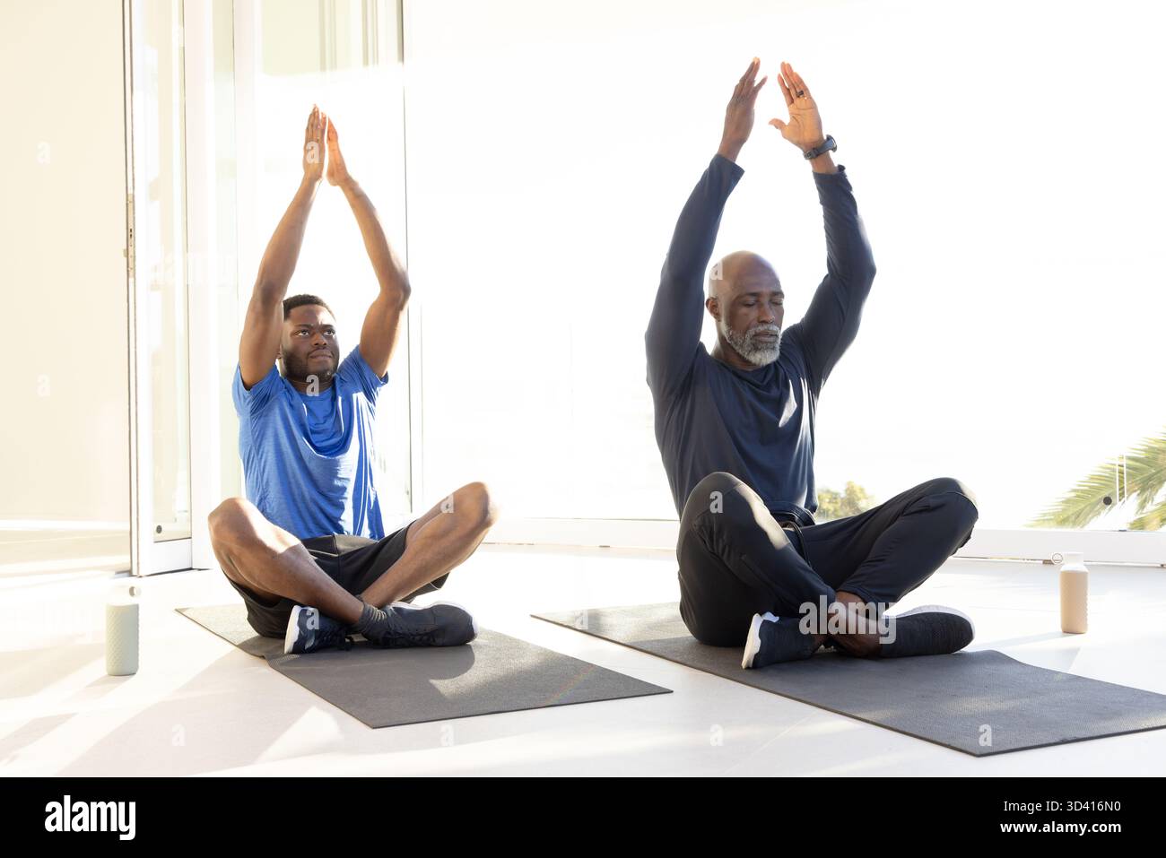 Étirer les partenaires de yoga afro-américains levant les bras dans la chambre, avec des tapis de yoga et des bouteilles d'eau Banque D'Images