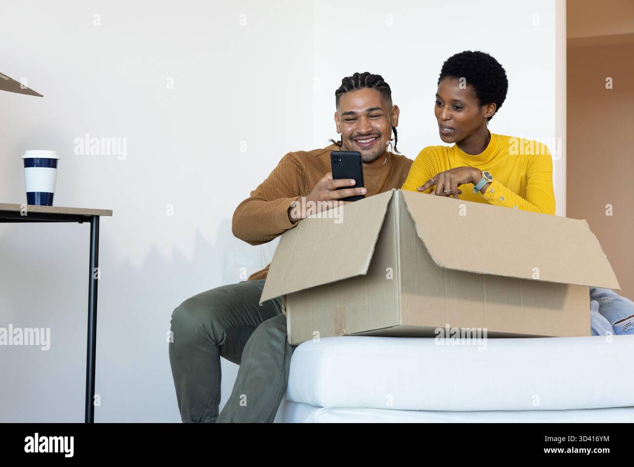 Déballage de divers couples assis sur le canapé dans le nouveau salon, avec boîte en carton et smartphone Banque D'Images