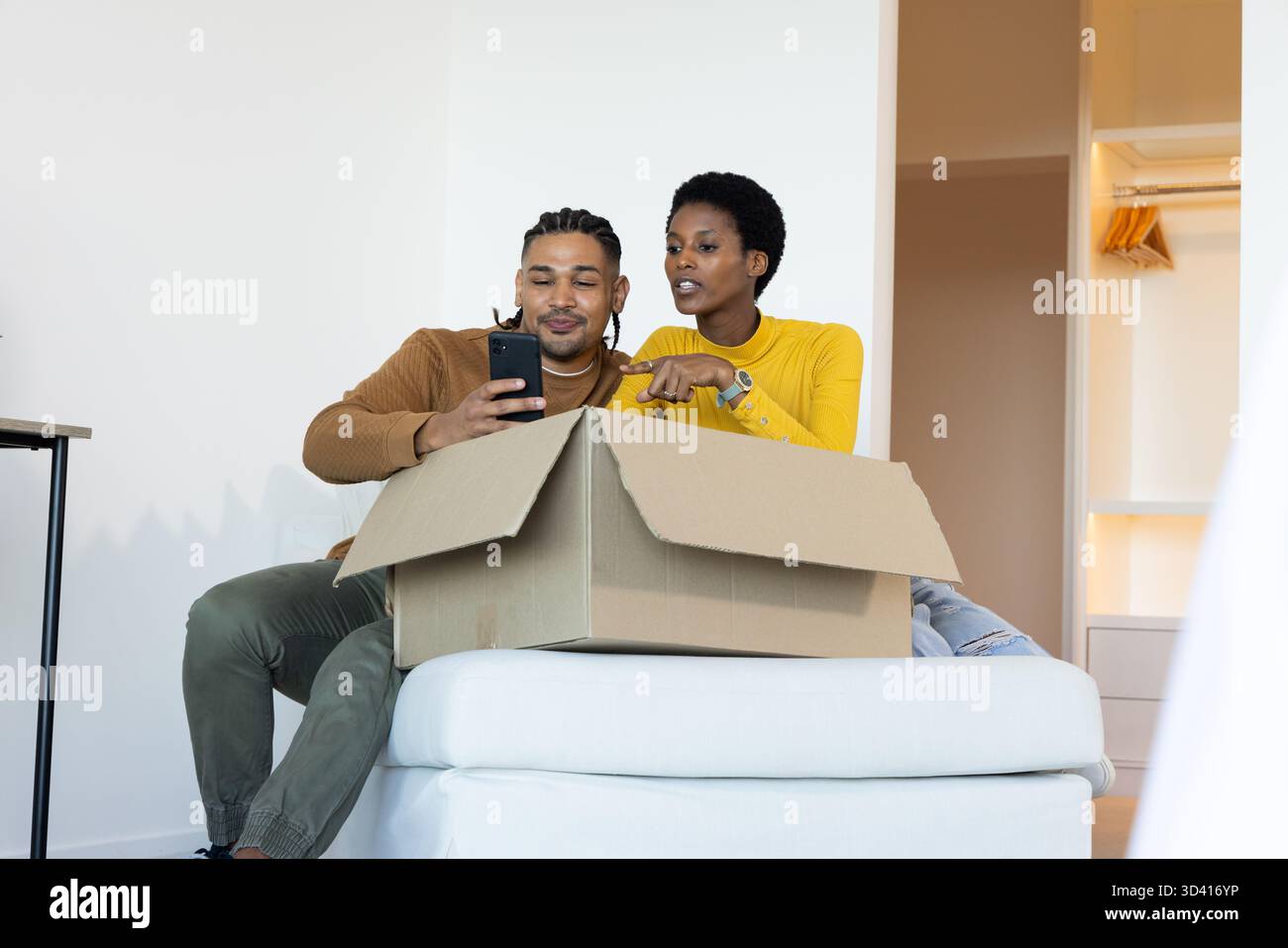 Couple diversifié déballage boîte en carton sur pouf rembourré dans la pièce clairsemée, vérification smartphone Banque D'Images
