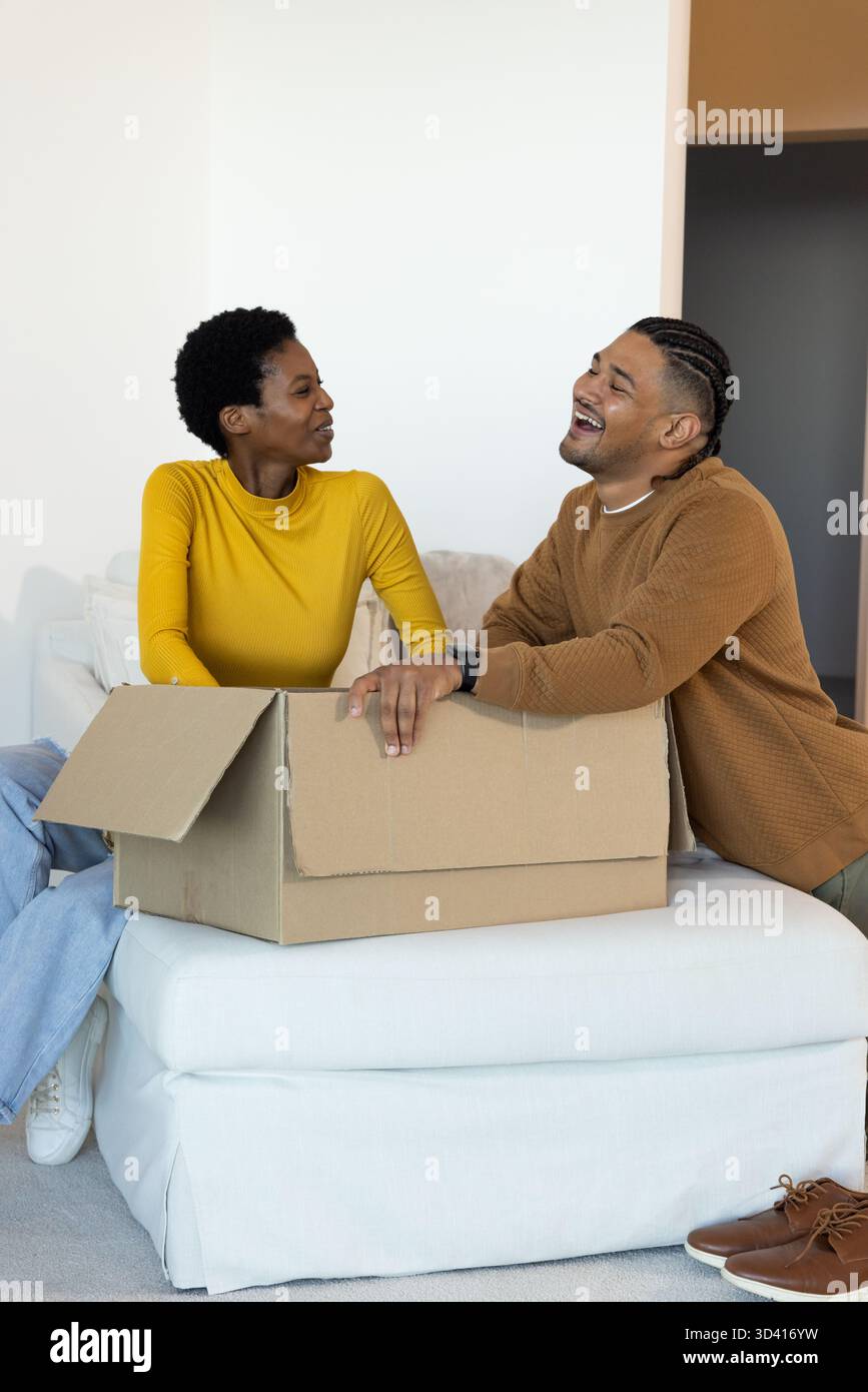 Rire couple diversifié assis sur un pouf blanc dans un salon lumineux, avec une boîte en carton mobile Banque D'Images