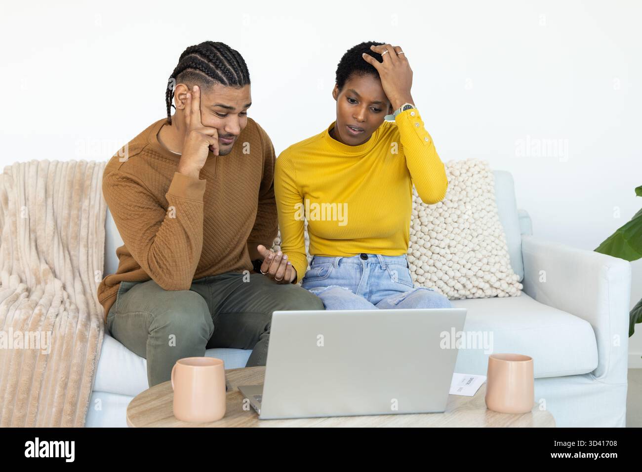 Regardant un couple diversifié assis sur un canapé dans le salon, avec des tasses pour ordinateur portable et plante Banque D'Images
