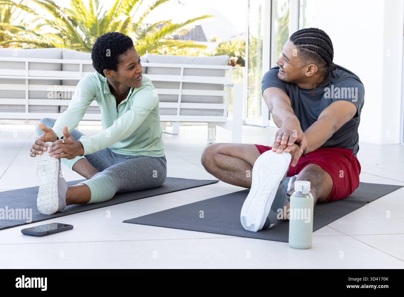 Couple diversifié s'étirant dans une salle d'entraînement lumineuse sur des tapis de yoga, avec smartphone et bouteille d'eau Banque D'Images