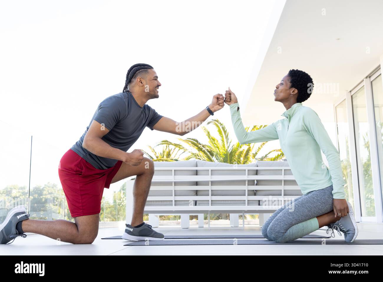 Agenouillement de divers amis se cognant les poings sur la terrasse extérieure, avec tapis d'exercice et balustrade en verre Banque D'Images