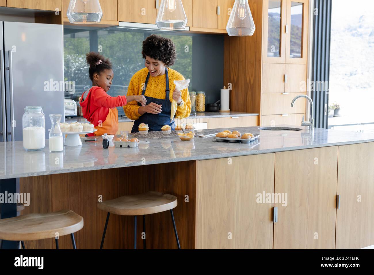 Duo mère-fille diversifié glaçant cupcakes à l'îlot de cuisine, avec sac à passepoil Banque D'Images