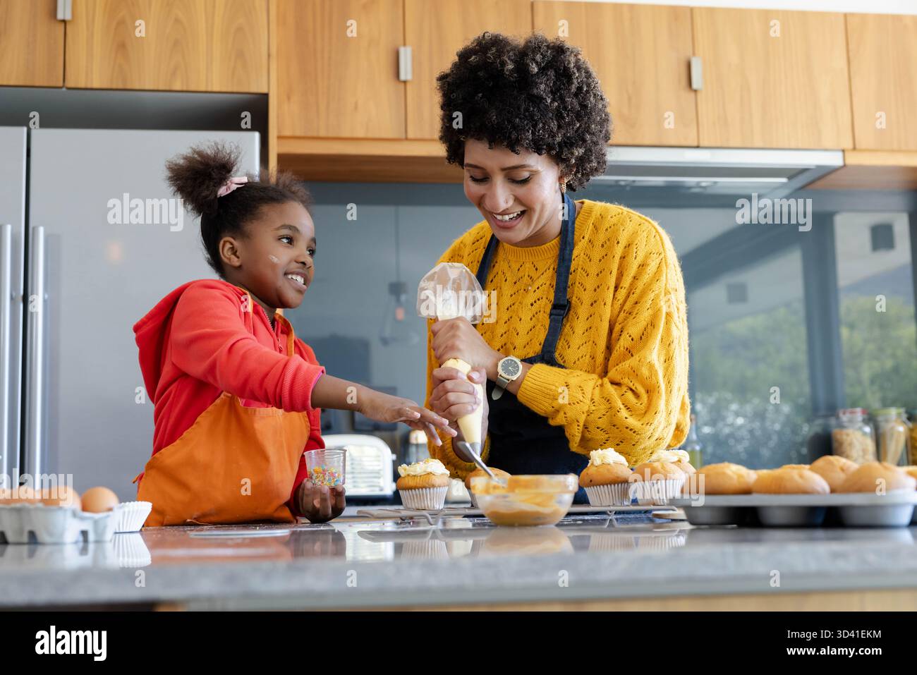 Mère et fille diversifiées décorant des cupcakes sur le comptoir dans la cuisine à la maison, avec des saupoudres Banque D'Images