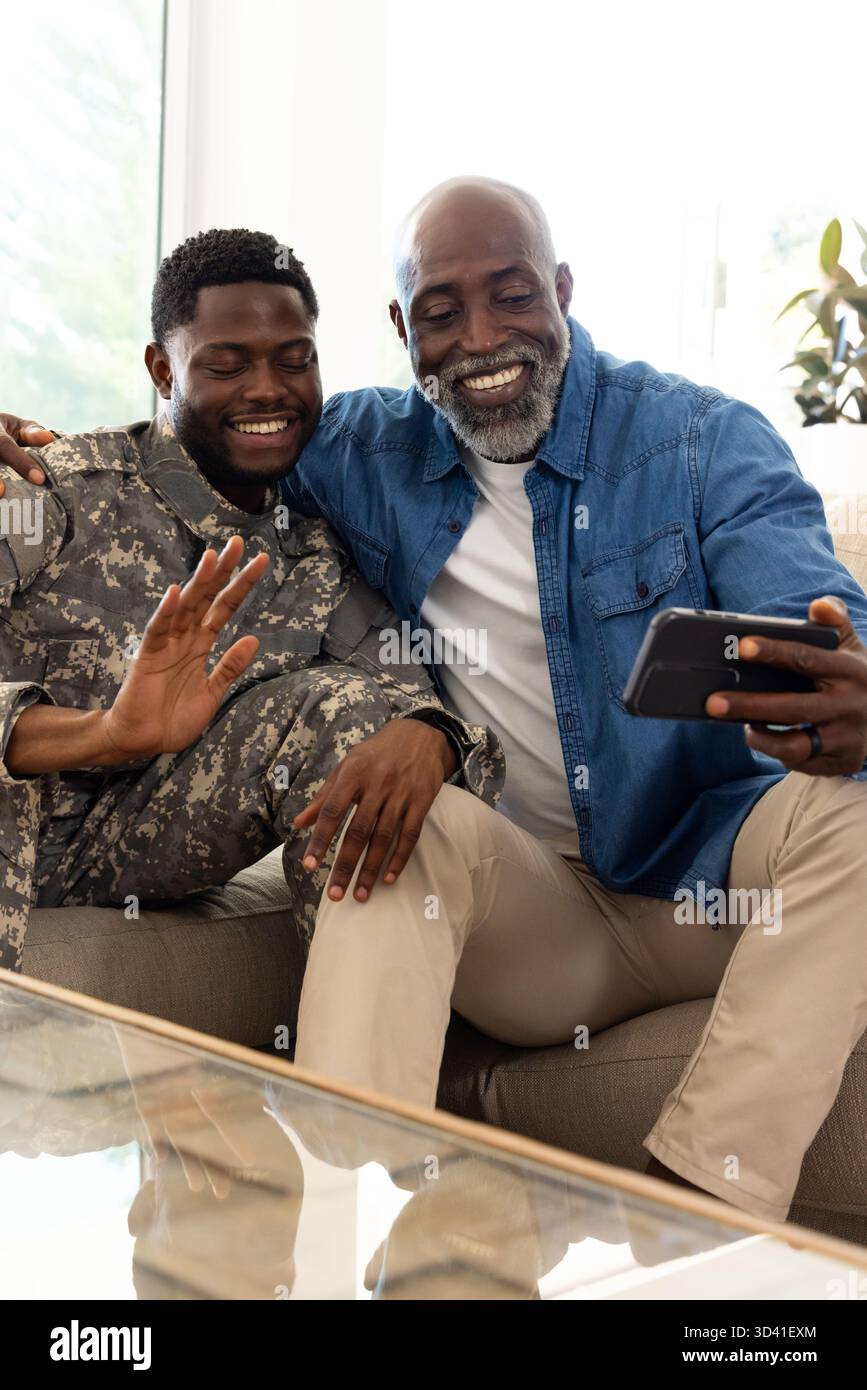 Sourire père afro-américain et fils partageant moment sur le canapé dans le salon, avec smartphone Banque D'Images