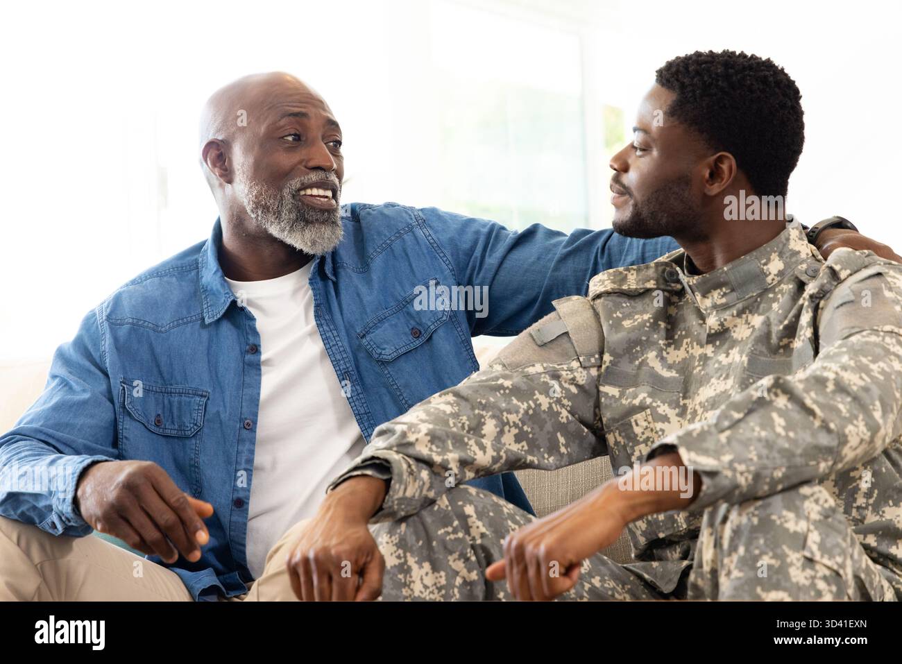 Père afro-américain et fils partageant la conversation sur le canapé à la maison, avec uniforme de camouflage Banque D'Images