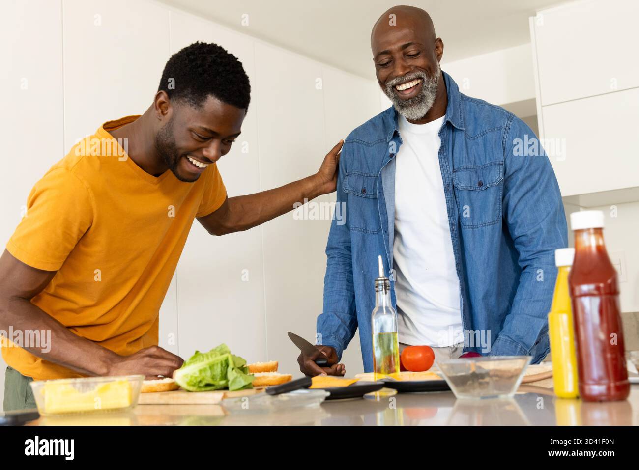 Père afro-américain et fils assemblant des hamburgers avec de la laitue dans la cuisine de la maison, avec planche à découper Banque D'Images