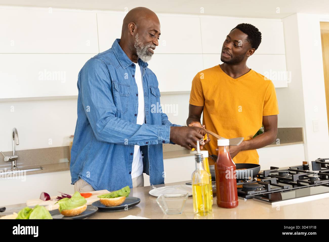 Père aîné afro-américain et fils cuisinant dans une cuisine moderne, avec spatule et petits pains à hamburger Banque D'Images