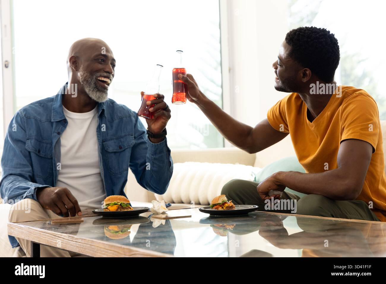 Clinking père afro-américain et fils assis sur le canapé dans le salon, avec thé glacé, hamburgers Banque D'Images