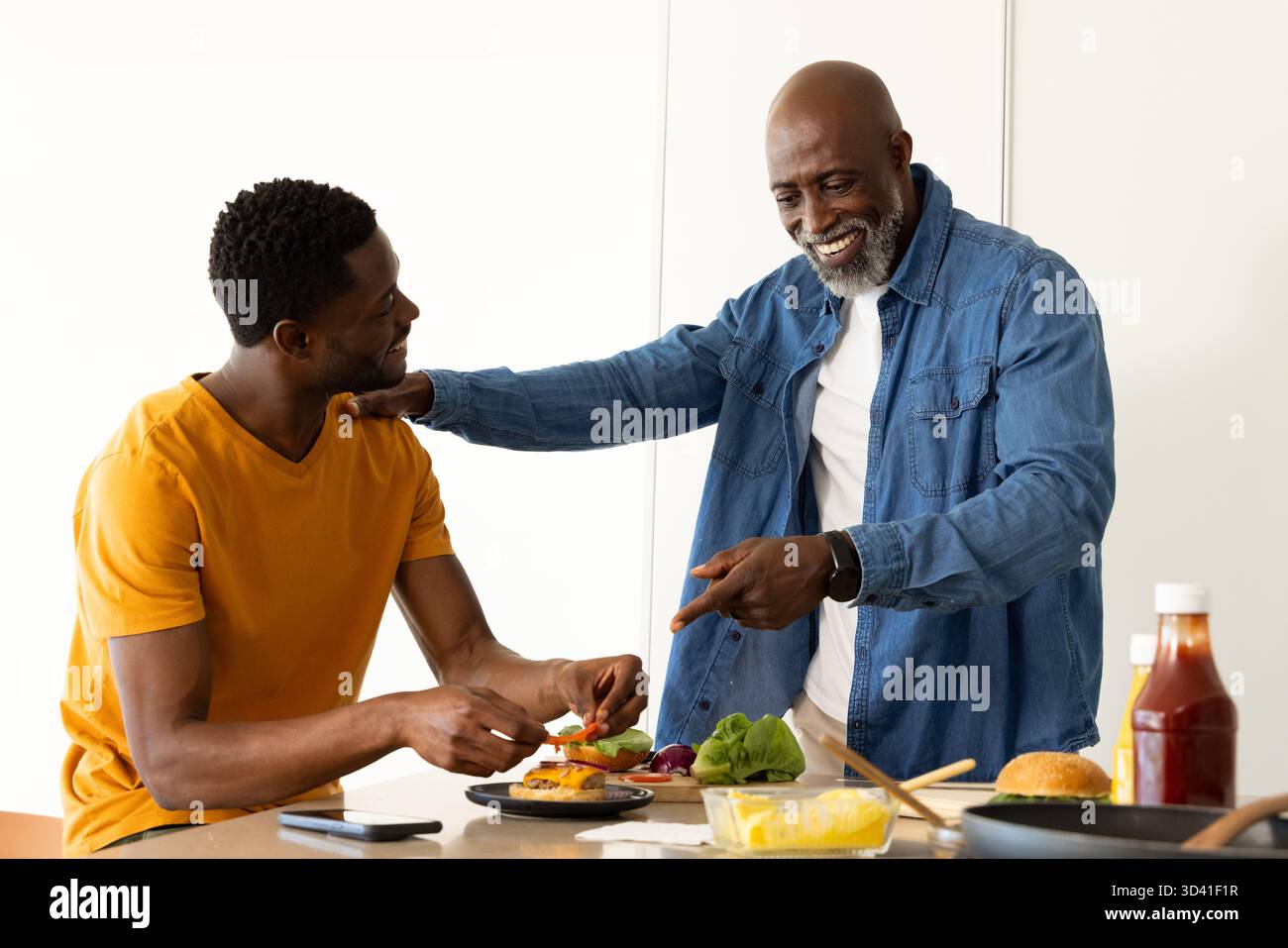 Père et fils afro-américains préparant des hamburgers dans une cuisine moderne, avec petits pains et smartphone Banque D'Images