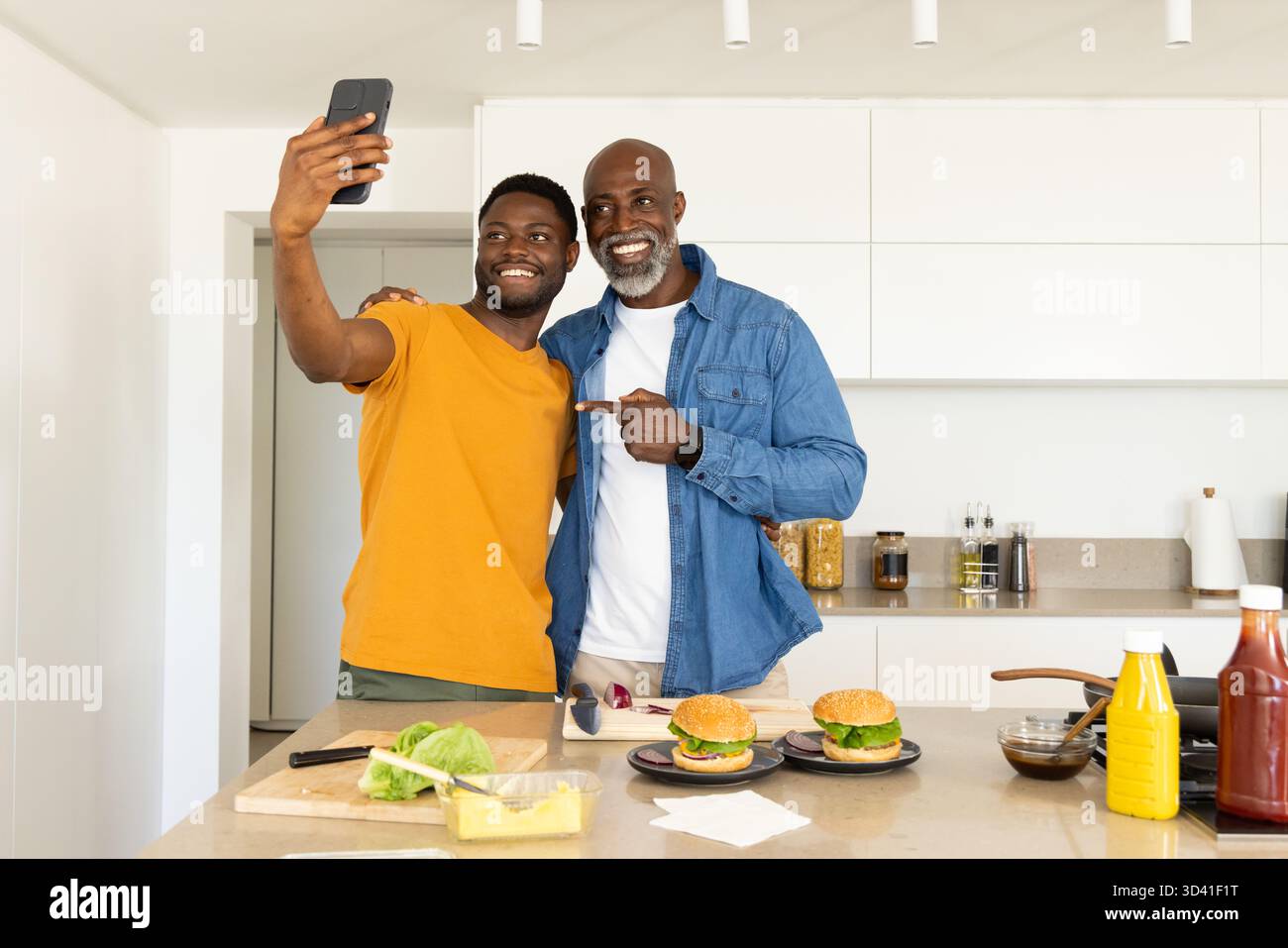 Posant père afro-américain et fils préparant des hamburgers à l'îlot de cuisine, avec smartphone Banque D'Images