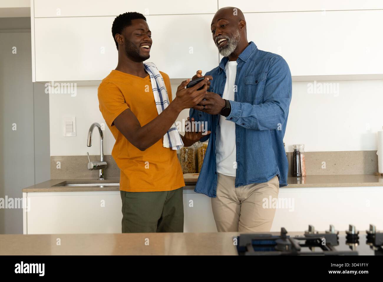 Broyage afro-américain père et fils à l'aide d'un moulin à poivre noir au comptoir de la cuisine, avec serviette Banque D'Images