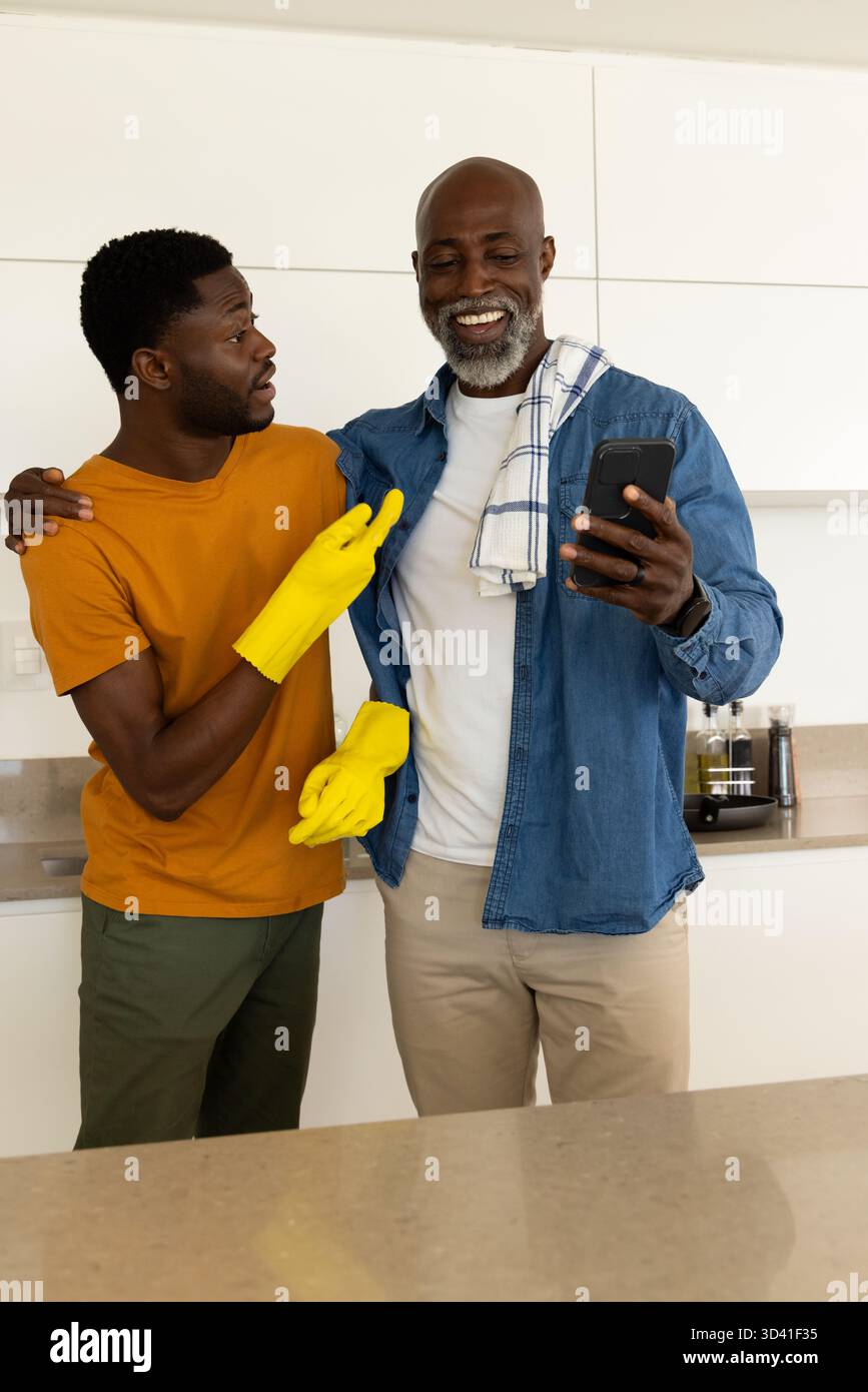 Sourire père afro-américain et fils regardant smartphone dans la cuisine moderne, avec des gants jaunes Banque D'Images