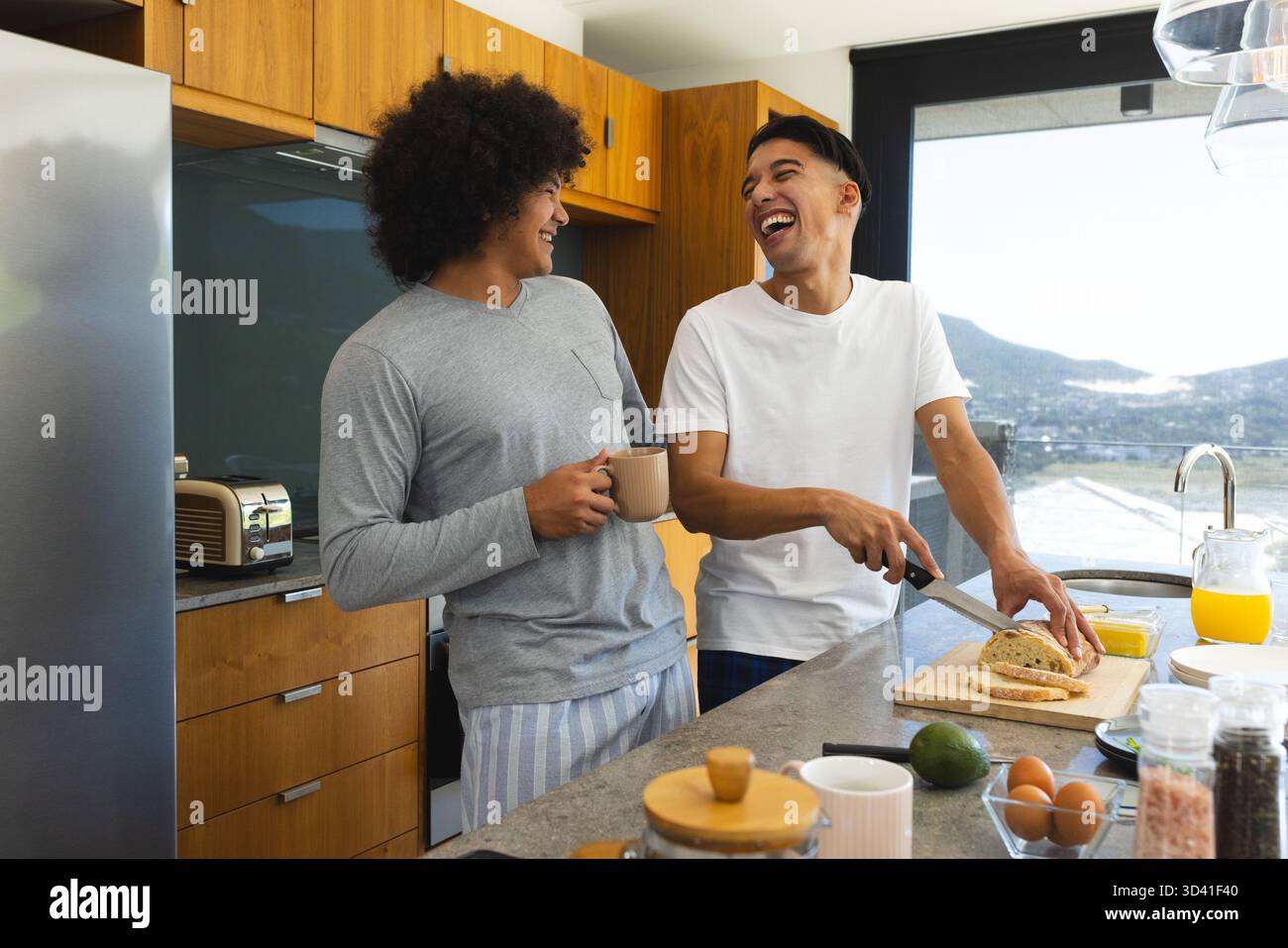 Partage du petit déjeuner, couple masculin diversifié tranchant du pain à l'îlot de cuisine, avec planche à découper et tasses Banque D'Images