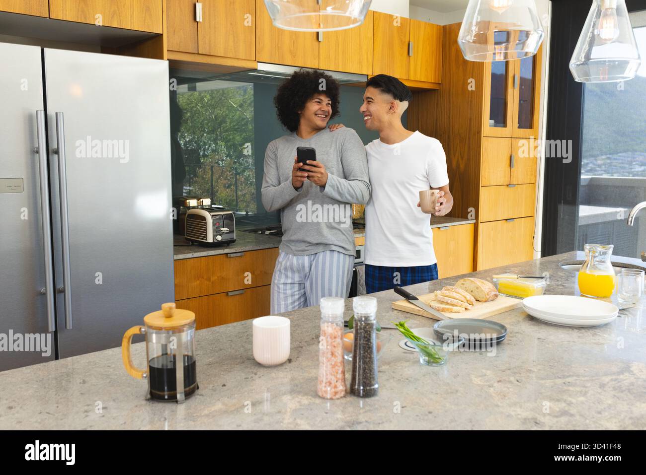 Souriant divers partenaires masculins debout à l'île de marbre dans la cuisine, avec smartphone et tasse à café Banque D'Images