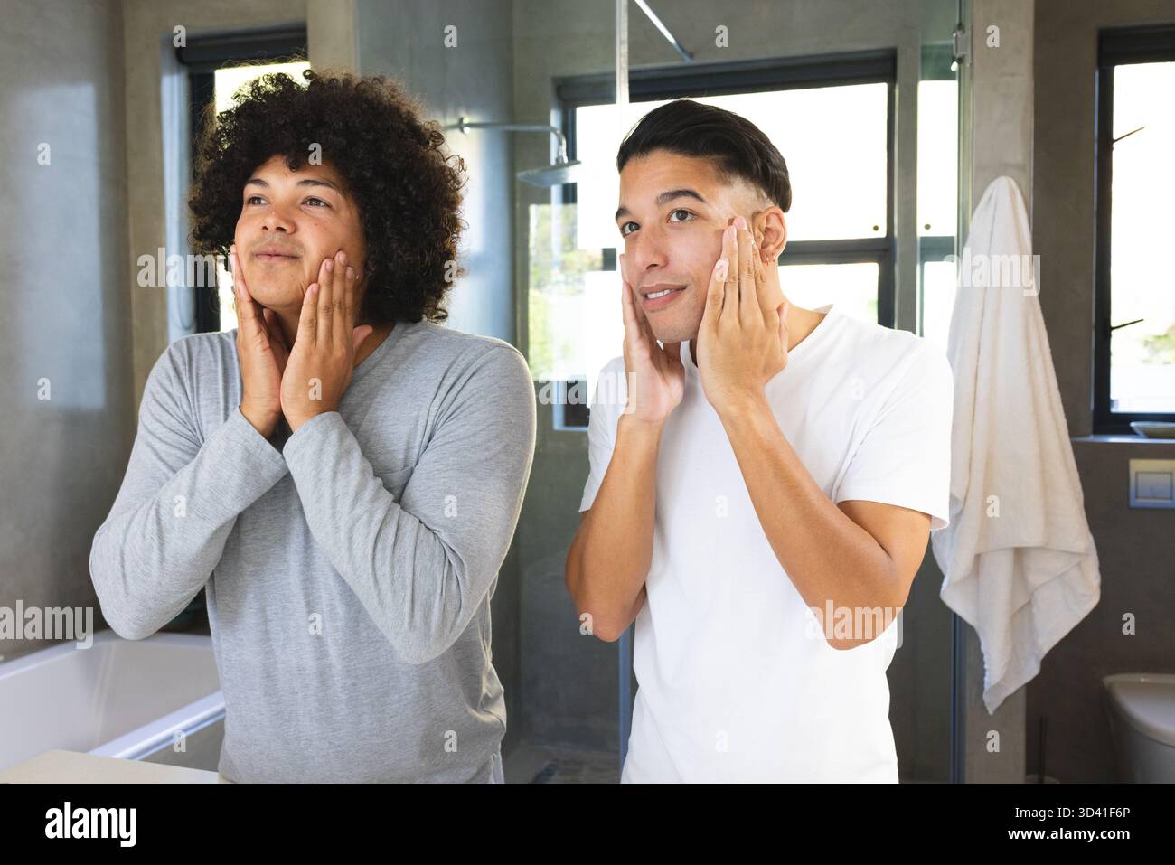 Application de crème de soin de la peau divers amis masculins debout à l'évier de salle de bain, avec grand miroir et serviette Banque D'Images