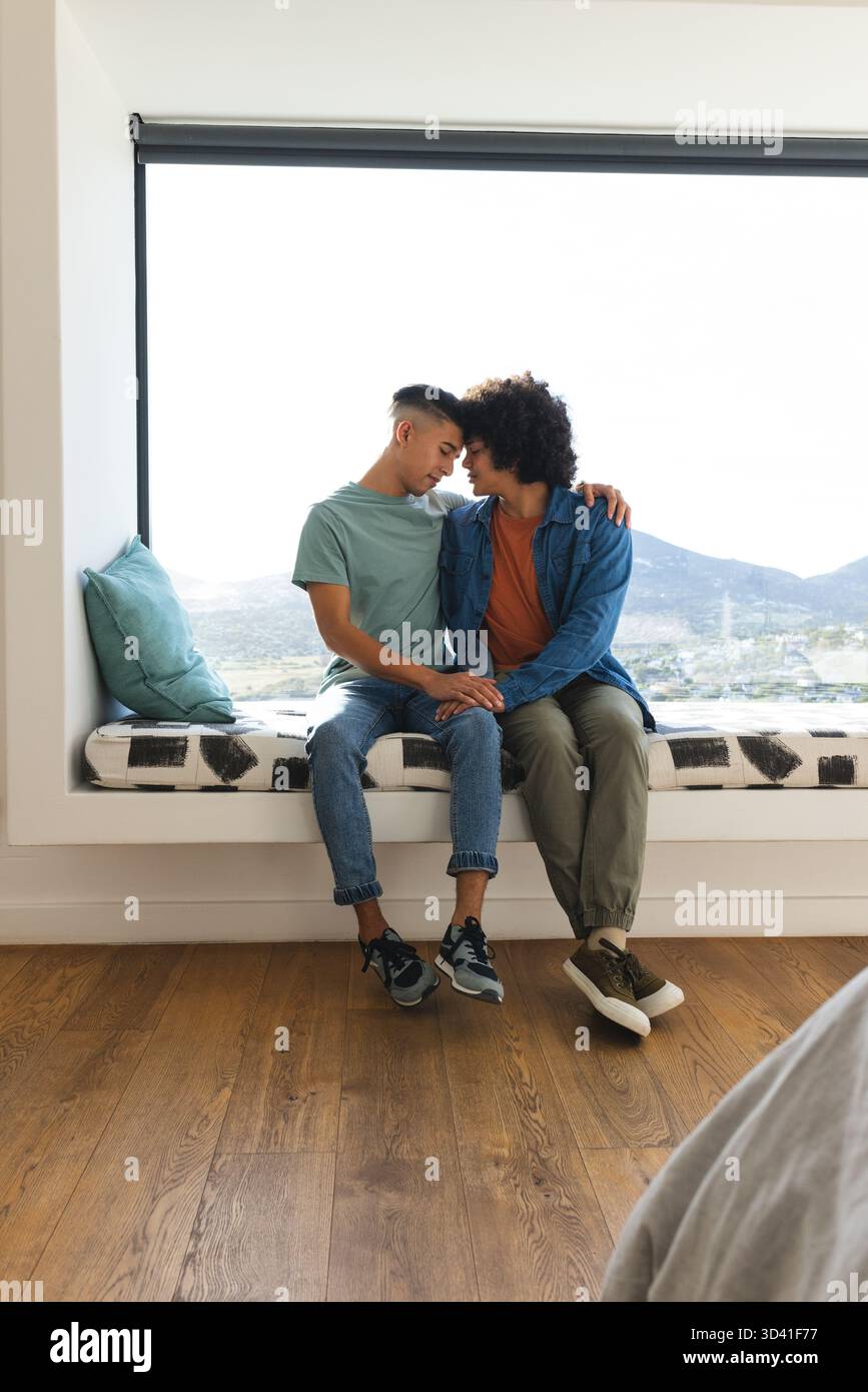 Penchant les têtes ensemble, couple masculin tenant les mains sur le siège de fenêtre par la grande fenêtre avec oreiller sarcelle Banque D'Images