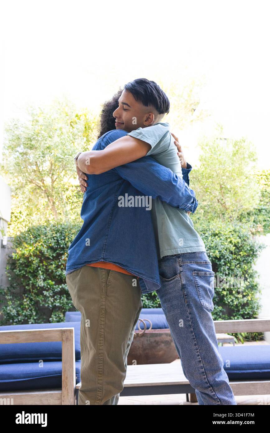 Embrassant père et fils adultes debout sur le patio arrière, avec table basse en bois et bancs bleus Banque D'Images