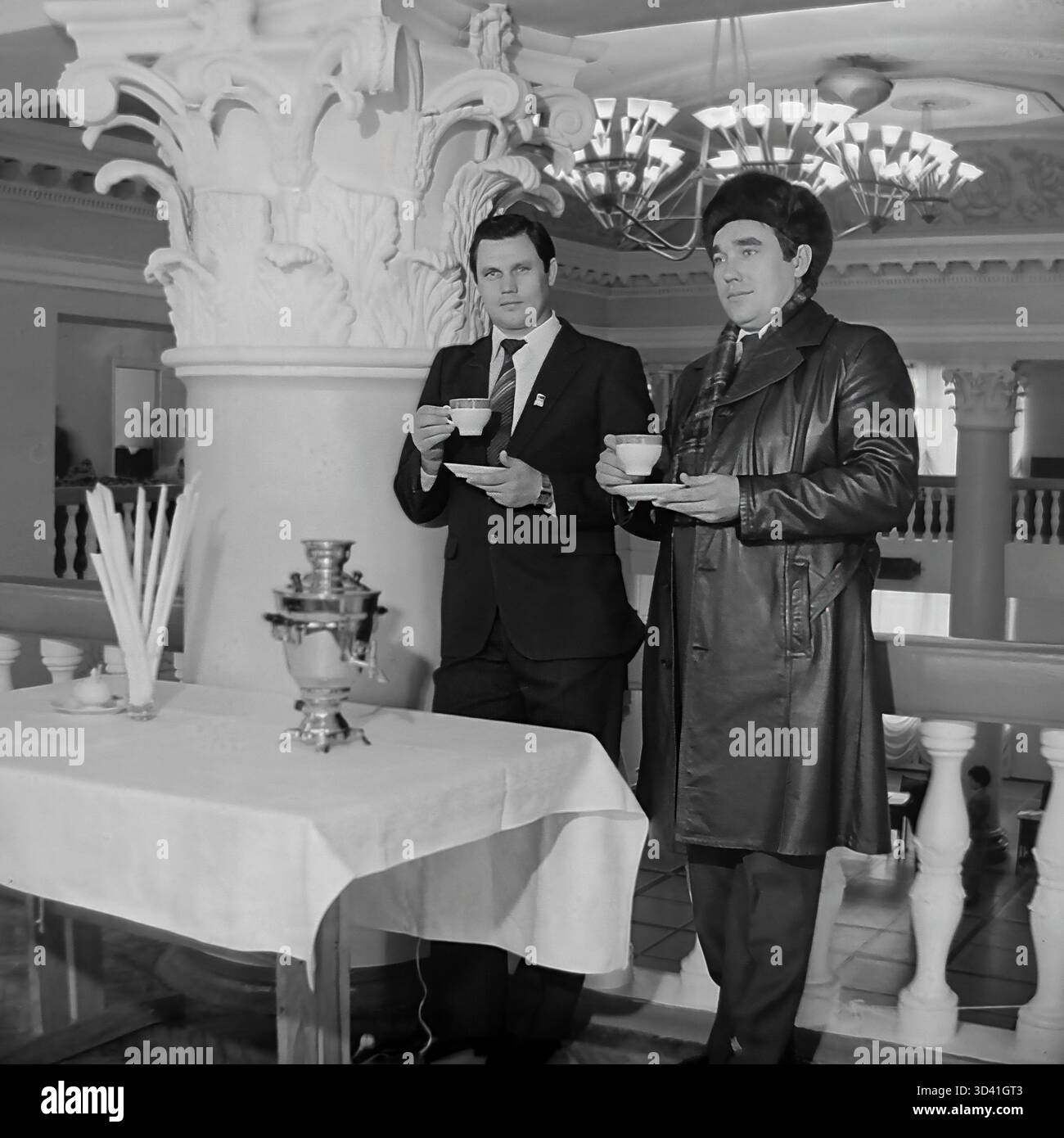 Une photo d'archive de 1984 montre deux délégués masculins à une conférence à la Maison de la culture Artyom (DK) à Sloviansk, RSS d'Ukraine. Les hommes sont en pause thé, debout près d'une table avec un samovar traditionnel. Ils sont dans le grand foyer, reconnaissable à ses colonnes ornées et ses lustres. L'homme à droite porte un chapeau de fourrure distinctif (ushanka), suggérant qu'il fait froid. Cela capture un moment de discussion lors d'un événement soviétique officiel. Cela montre la vie quotidienne dans le Donbass paisible avant la guerre. Banque D'Images