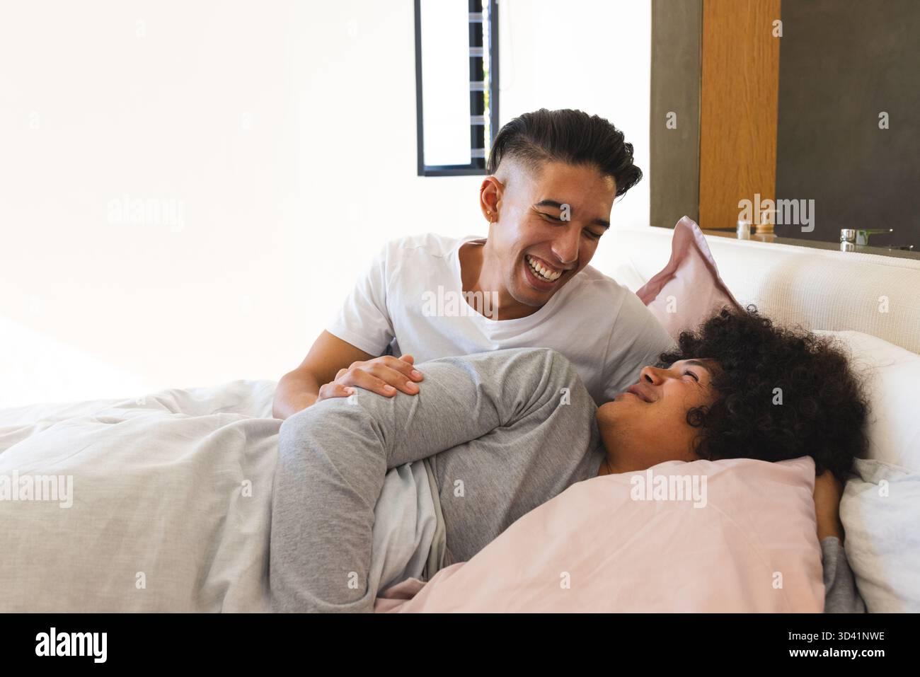 Rire divers partenaires masculins allongés sur le lit dans une chambre moderne, avec une couverture grise et des oreillers roses Banque D'Images