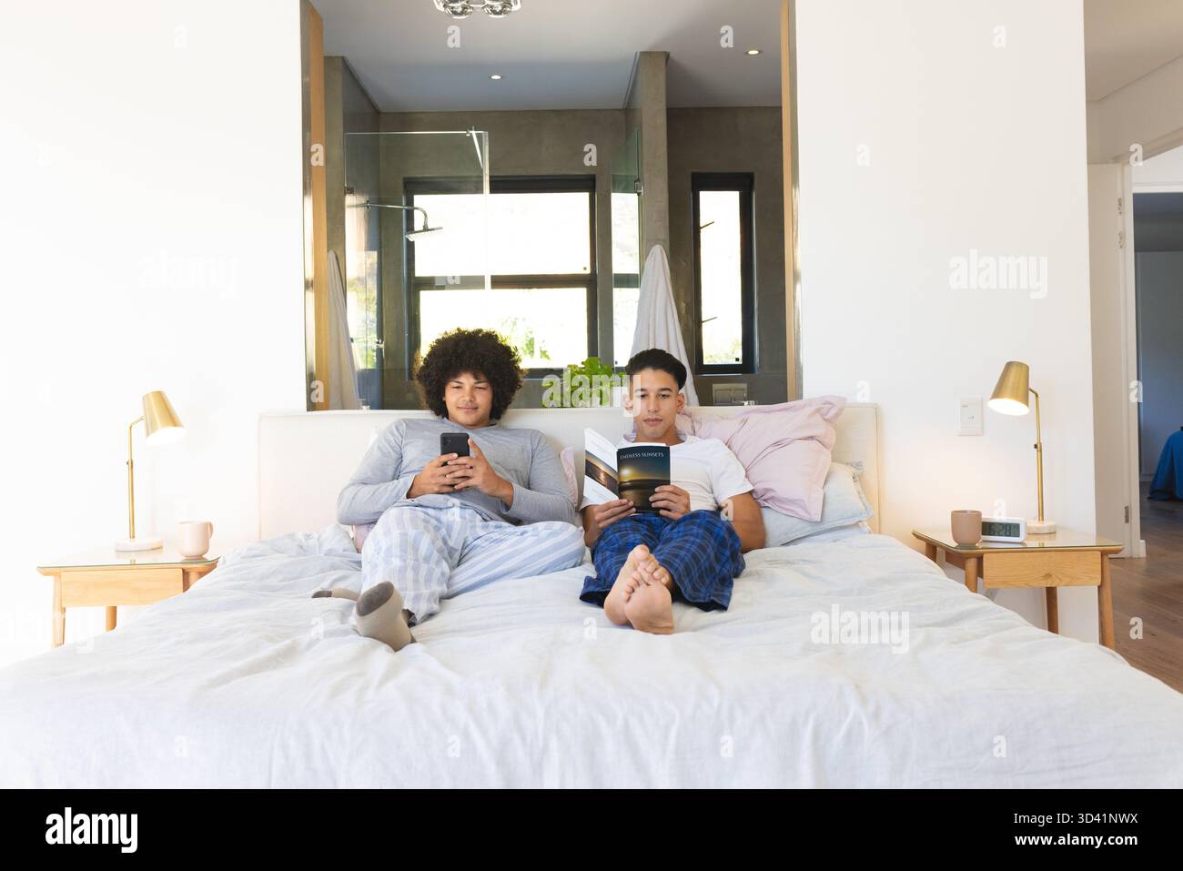 Relaxant couple masculin diversifié à l'aide d'un smartphone et d'un livre de lecture sur le lit, avec des lampes, un espace de copie Banque D'Images