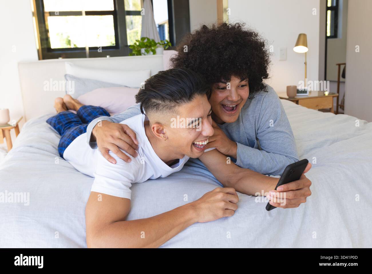 Rire partenaires masculins couchés sur le lit dans une chambre lumineuse, tenant smartphone près de la table de chevet Banque D'Images